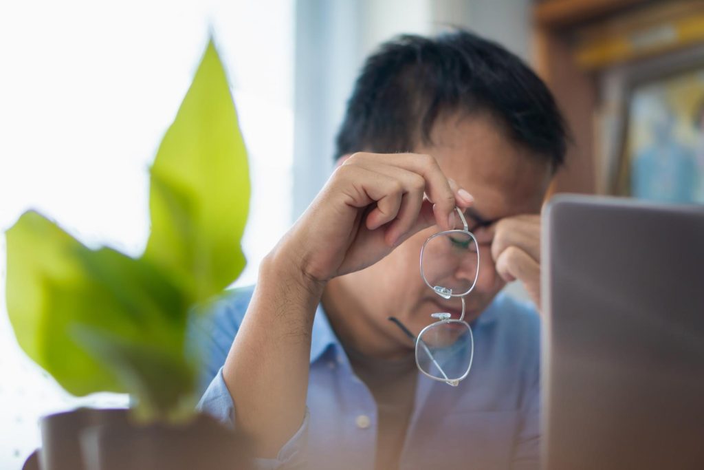Stressed man removing glasses and rubbing his eyes while working on a laptop, highlighting the need for presbyopia treatment.