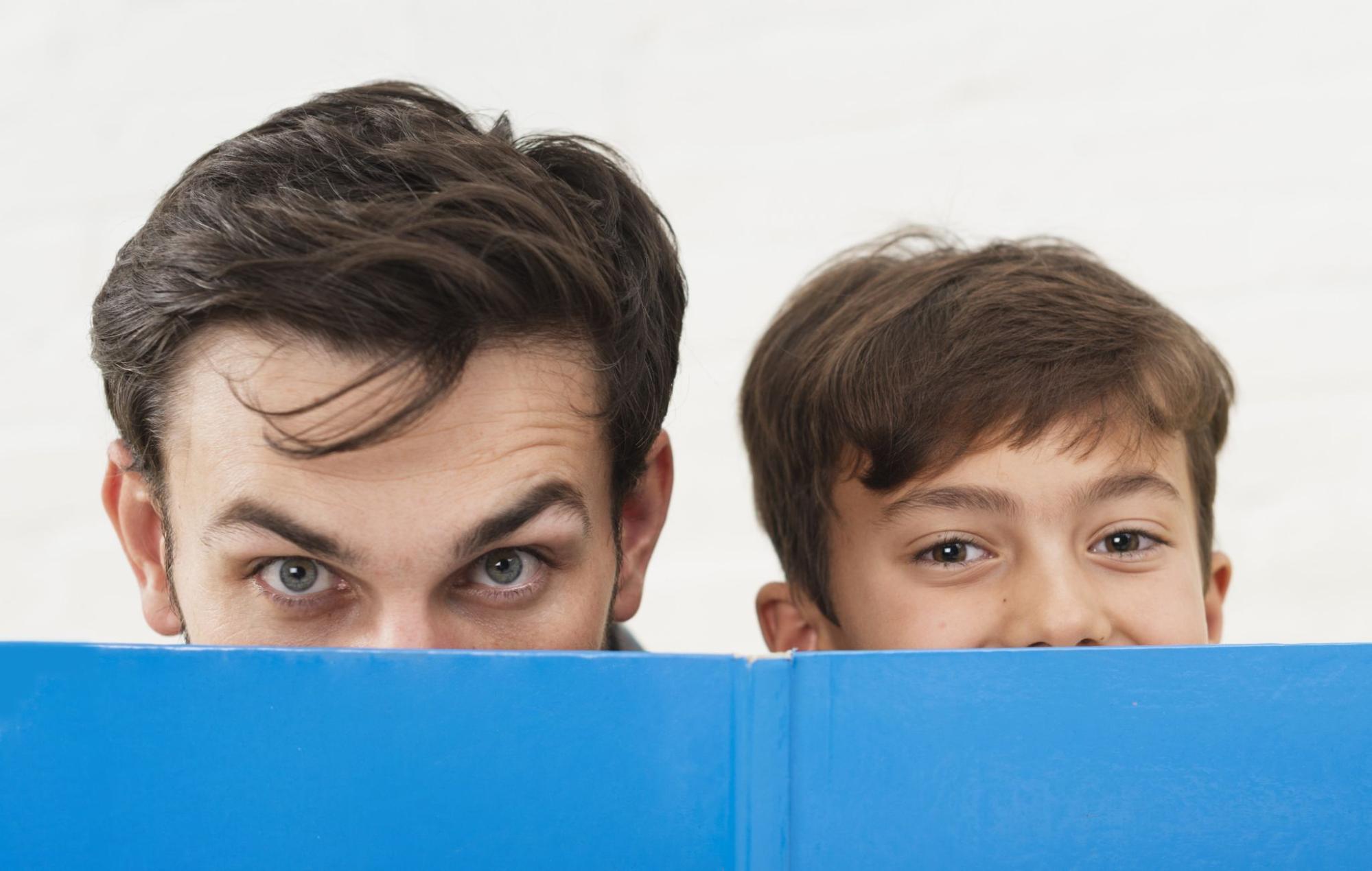 Father and son peeking playfully over book, symbolizing early detection and amblyopia lazy eye treatment for adults