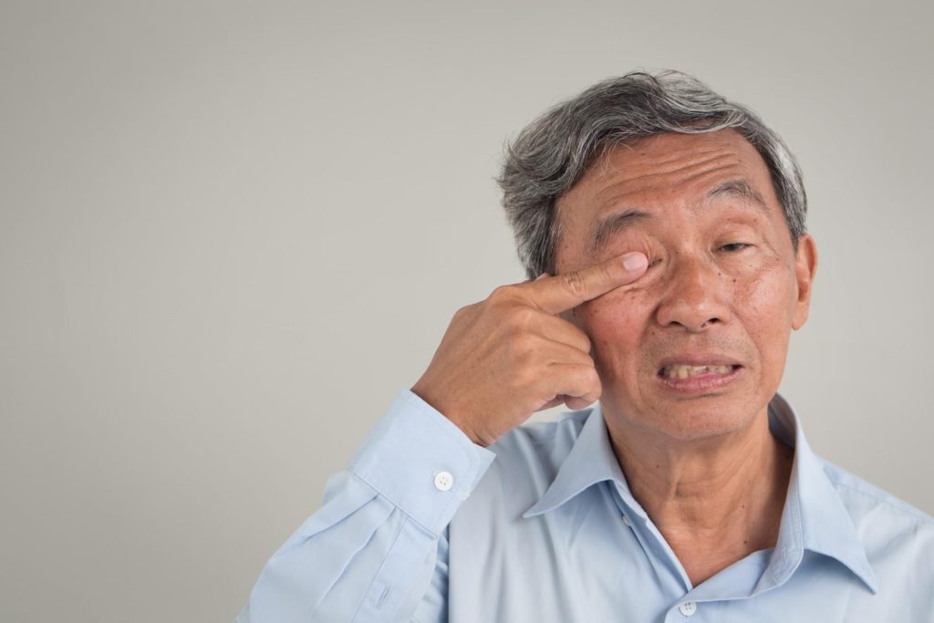 Elderly man pointing to his eyelid, possibly indicating ptosis of eyelid and illustrating potential ptosis causes in older adults.
