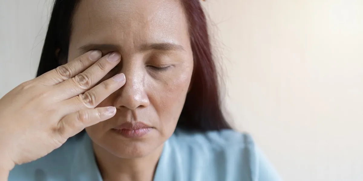 Woman in blue shirt touching her eye with closed eyes and pained expression