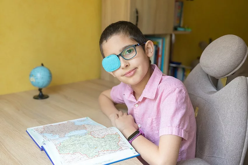 A boy wearing glasses with an eye patch sits at a desk with a map book and small globe, smiling at the camera