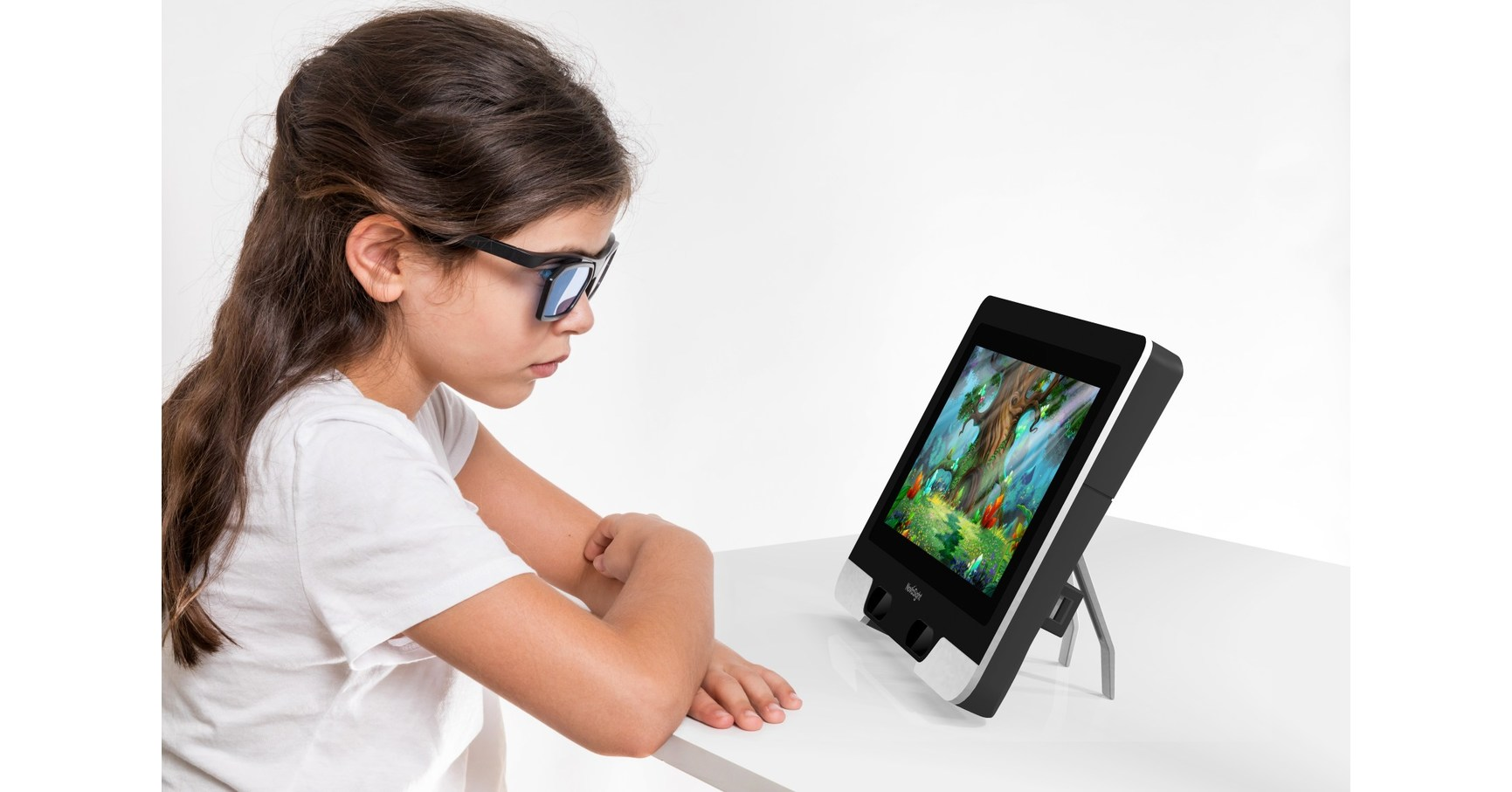 A girl wearing 3D glasses looks at a colorful forest scene on a small tablet-like device used for vision therapy