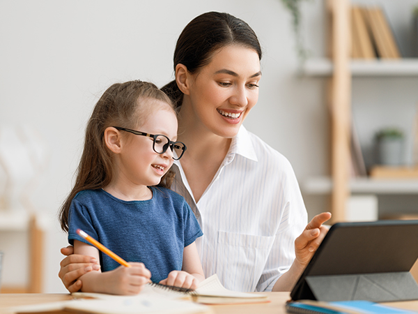 A mother smiles while helping her young daughter, who wears glasses, with schoolwork at a desk with a tablet and notebooks