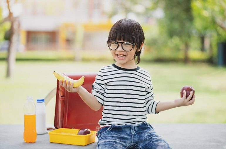 A smiling boy wearing glasses holds a banana and an apple, with a healthy lunch and drinks in front of him outdoors