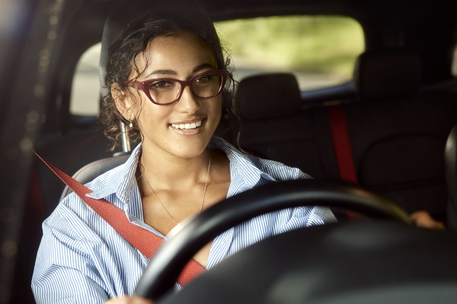 A smiling woman wearing glasses and a seatbelt drives a car, hands on the steering wheel.
