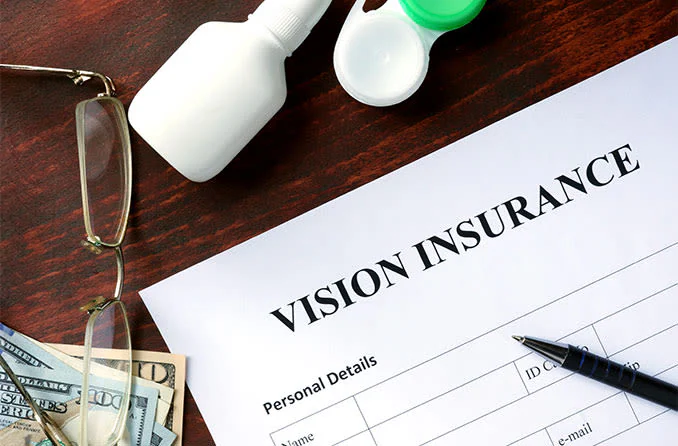 A vision insurance form sits on a desk with glasses, eye drops, a contact lens case, cash, and a pen