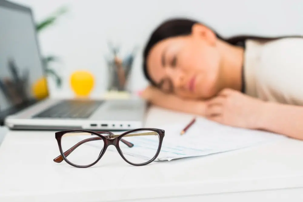 A woman falls asleep at her desk next to a laptop and papers, with her glasses set down in front of her
