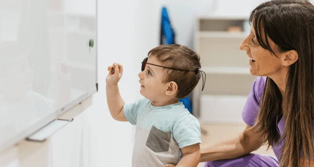 A woman holds a young boy wearing an eye patch as he reaches up to touch a whiteboard