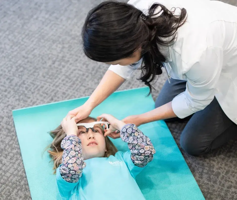 A woman in a lab coat helps fit tracking glasses on a girl lying on a mat