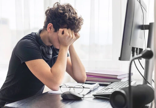 A young man sits stressed at a desk with his head in his hands, glasses removed, in front of a computer and papers
