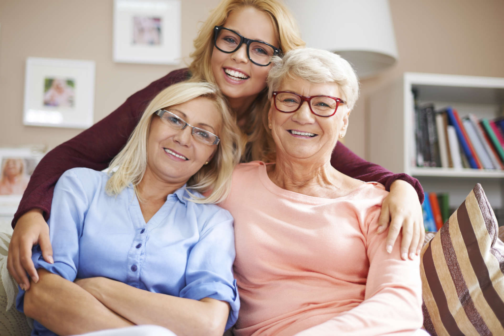 Three generations of women, all wearing glasses, smile together on a couch