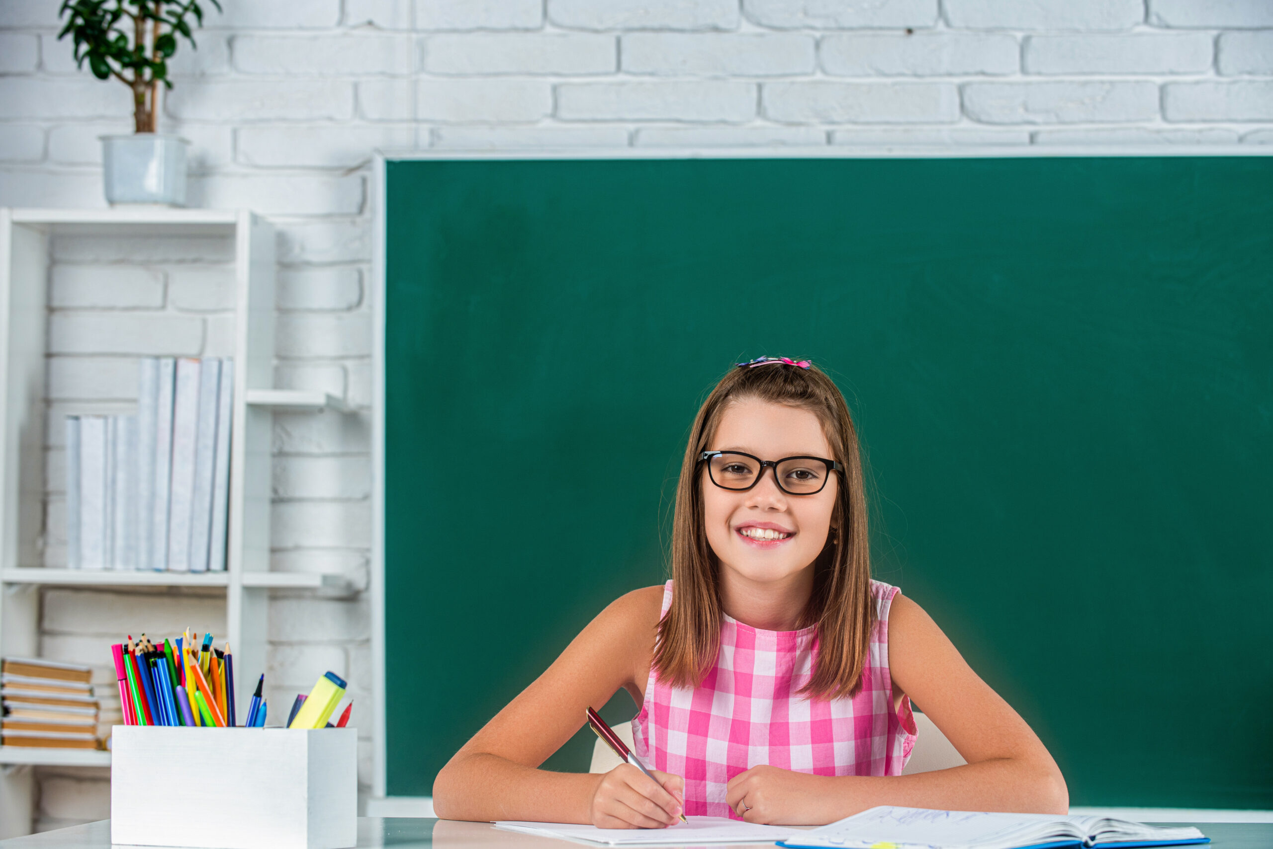 A smiling girl wearing glasses sits at a desk in front of a green chalkboard, with a bookshelf and pencil cup beside her