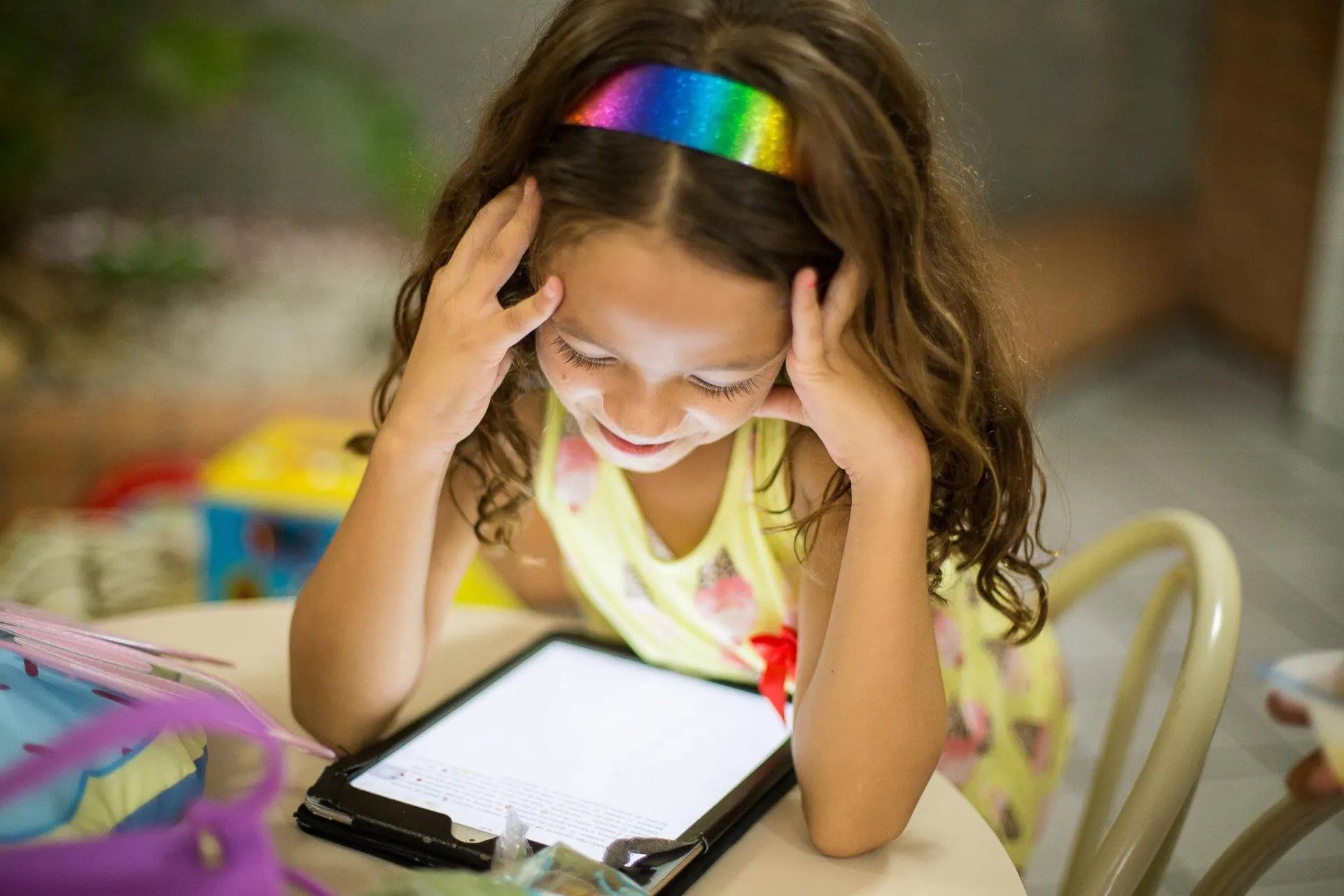 A smiling girl wearing a rainbow headband holds her head in her hands while looking down at a tablet