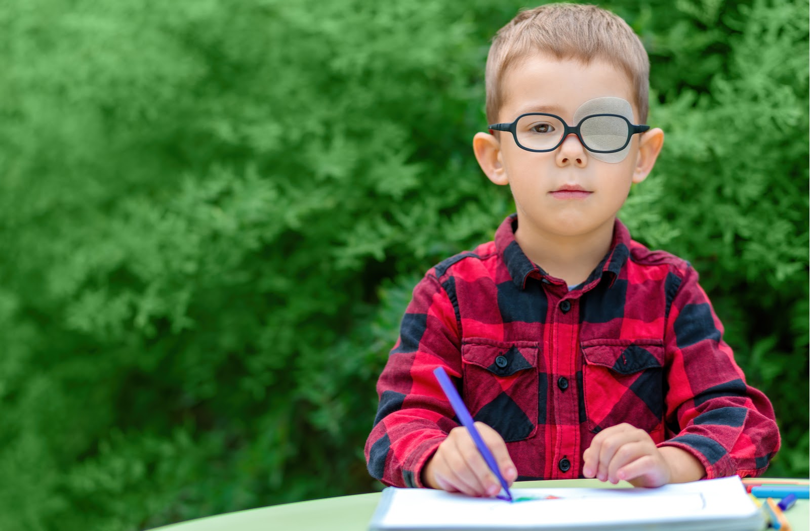 A boy wearing glasses with an eye patch stands outdoors in front of greenery, holding a pencil