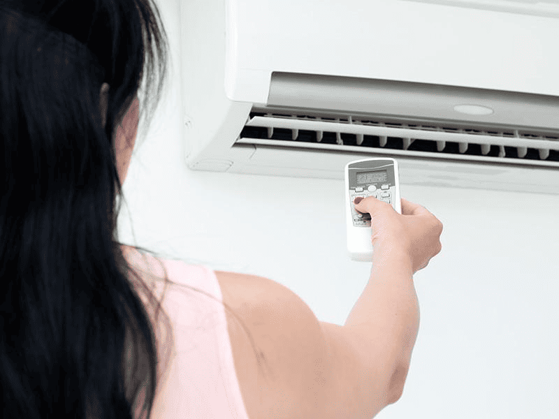 A woman adjusting the air conditioning remote control under an indoor AC unit