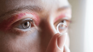 A person holding a contact lens close to their eye, with visible redness around the eyelids