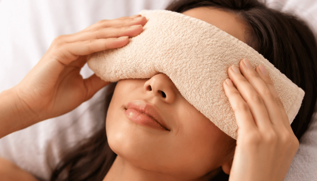 Woman applying a warm beige compress over both closed eyes.