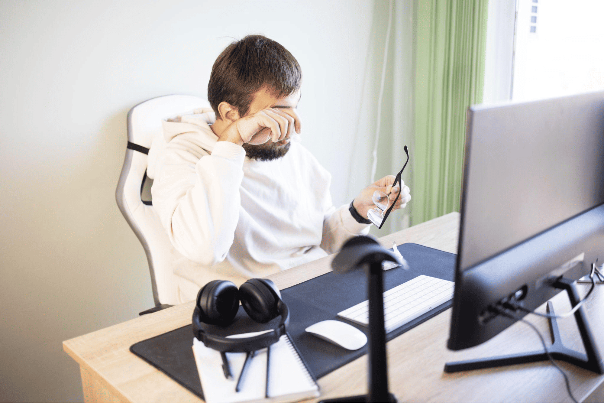 Person at desk in front of computer rubbing eyes with hand, holding glasses