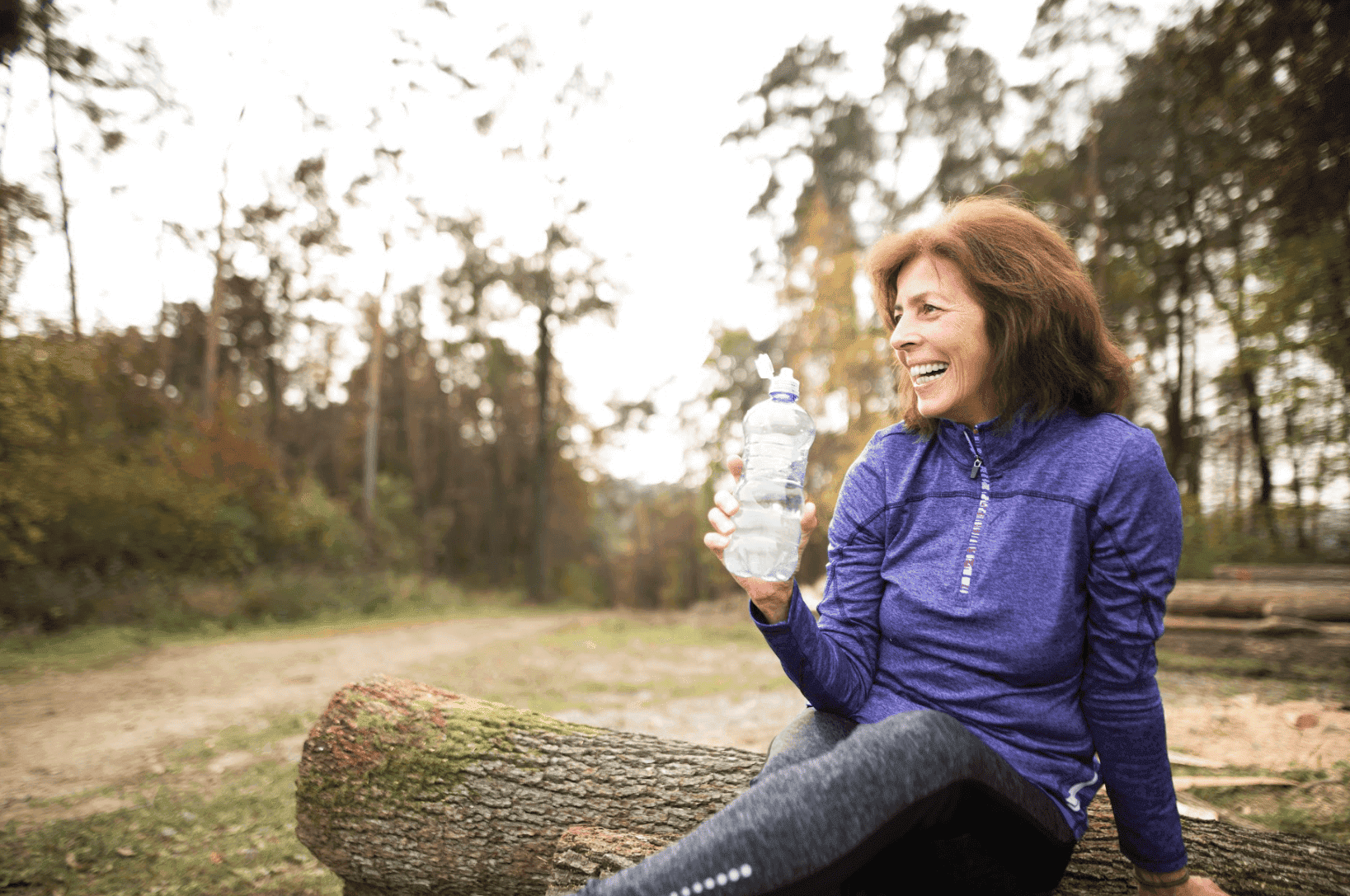 Woman in purple athletic jacket sitting on a log in forest, holding water bottle and smiling