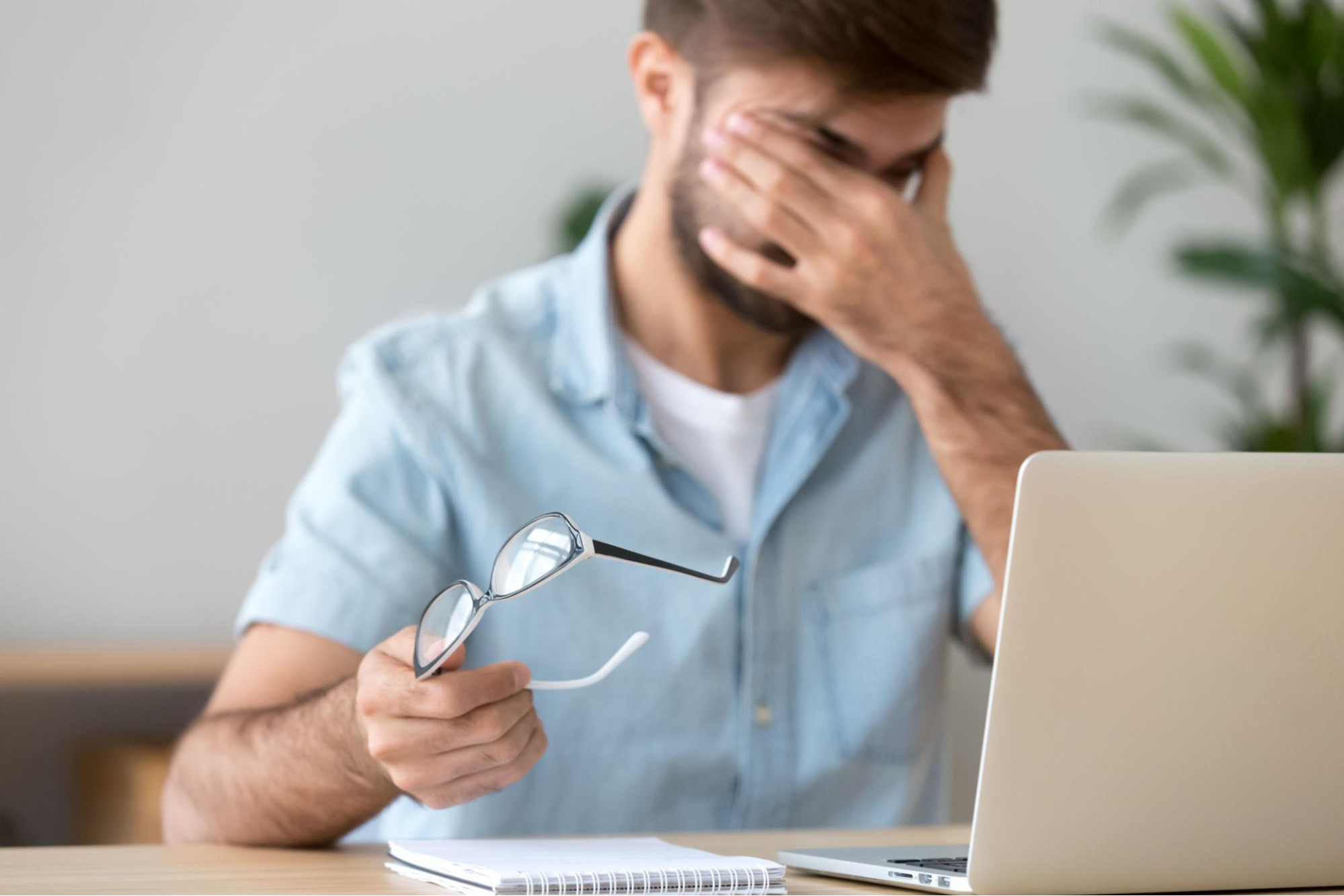 A man rubbing his tired eyes while holding his glasses in front of his laptop