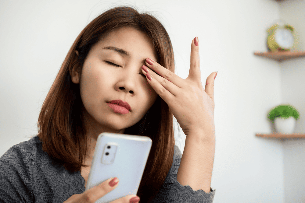 A woman rubbing her eye while looking at her phone